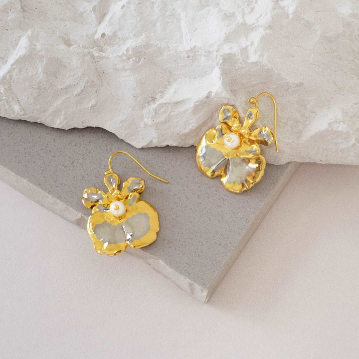 Risis Oncidium Coco Drop Earrings (PG) - Gold-tone flower-shaped earrings with reflective surfaces and central pearl-like embellishments are displayed on a geometric stone stand.