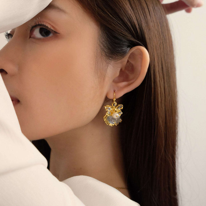 Risis Oncidium Coco Drop Earrings (PG) - A gold-toned earring with intricate floral design and a central pearl detail dangles from a woman's earlobe, complementing her elegant profile.