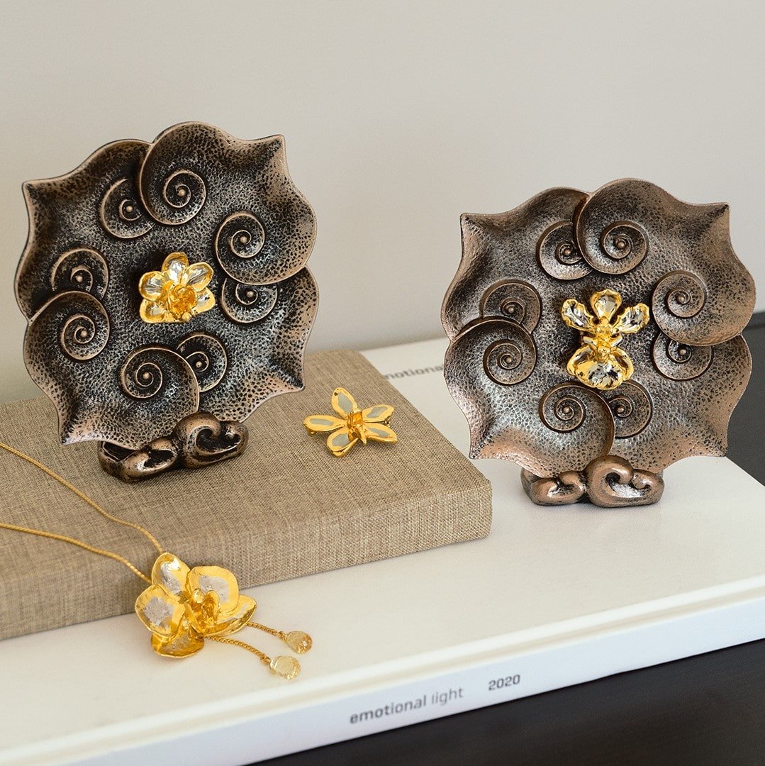 Risis Oncidium Mary Saporita Ruyi Plate - Two ornamental objects with flower and spiral motifs in bronze tones, accented with a central golden flower detail, accompanied by two subtle gold jewelry pieces.
