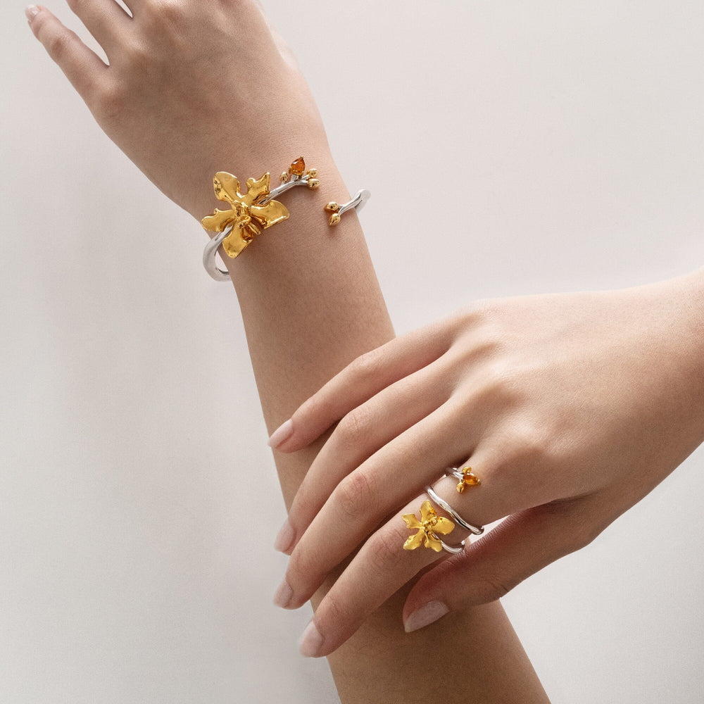 Risis Orchid Spiral Bangle - A silver ring adorned with a large golden orchid and smaller golden blooms, accented with bright amber-hued stones against a neutral surface.