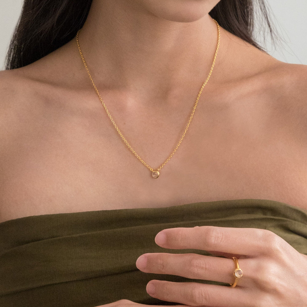 Risis Peranakan Jewel Necklace with Moonstone - A person is wearing a delicate gold necklace with a small, single gemstone pendant, complemented by a matching ring featuring a similar gem on an adjacent finger.