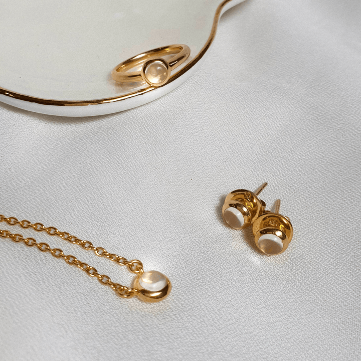Risis Peranakan Jewel Necklace with Moonstone - Golden jewelry with creamy beads: a pendant necklace, a ring, and a pair of stud earrings set on a textured white surface.