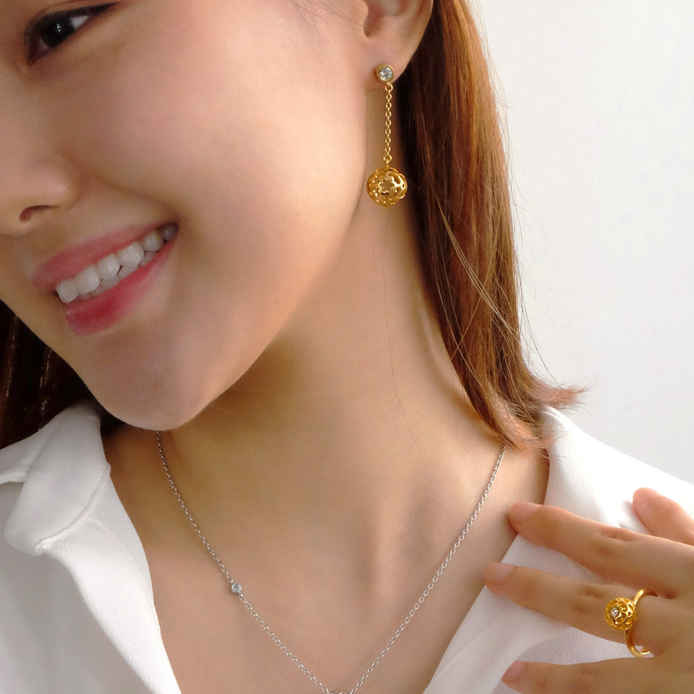 Risis Peranakan Spheres Dangling Earrings with White Topaz (G) - A smiling person wears a gold dangle earring with a filigree sphere, a delicate necklace with a small gemstone, and a matching intricate gold ring on their finger.