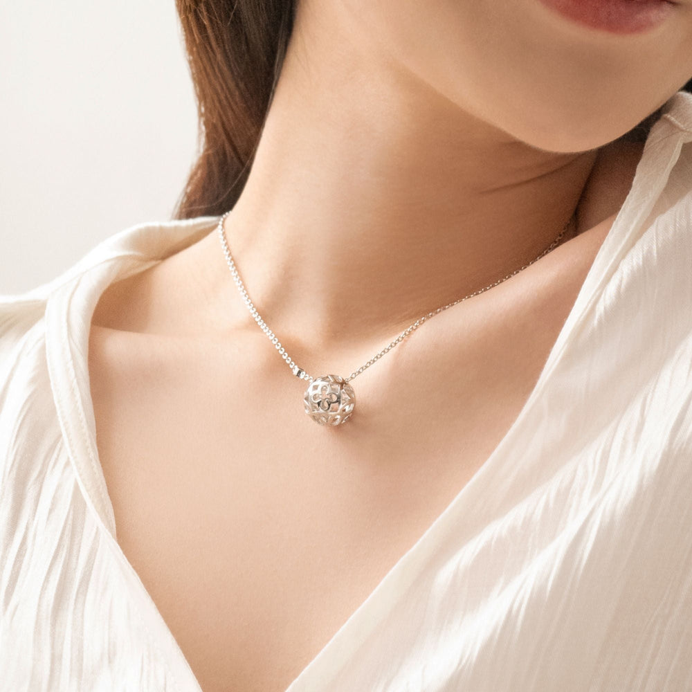 Risis Peranakan Spheres Large Necklace with Blue Topaz (RH) - An elegant silver pendant with a floral cut-out design dangles from a delicate chain, resting slightly on a person's collarbone against a soft, white fabric.