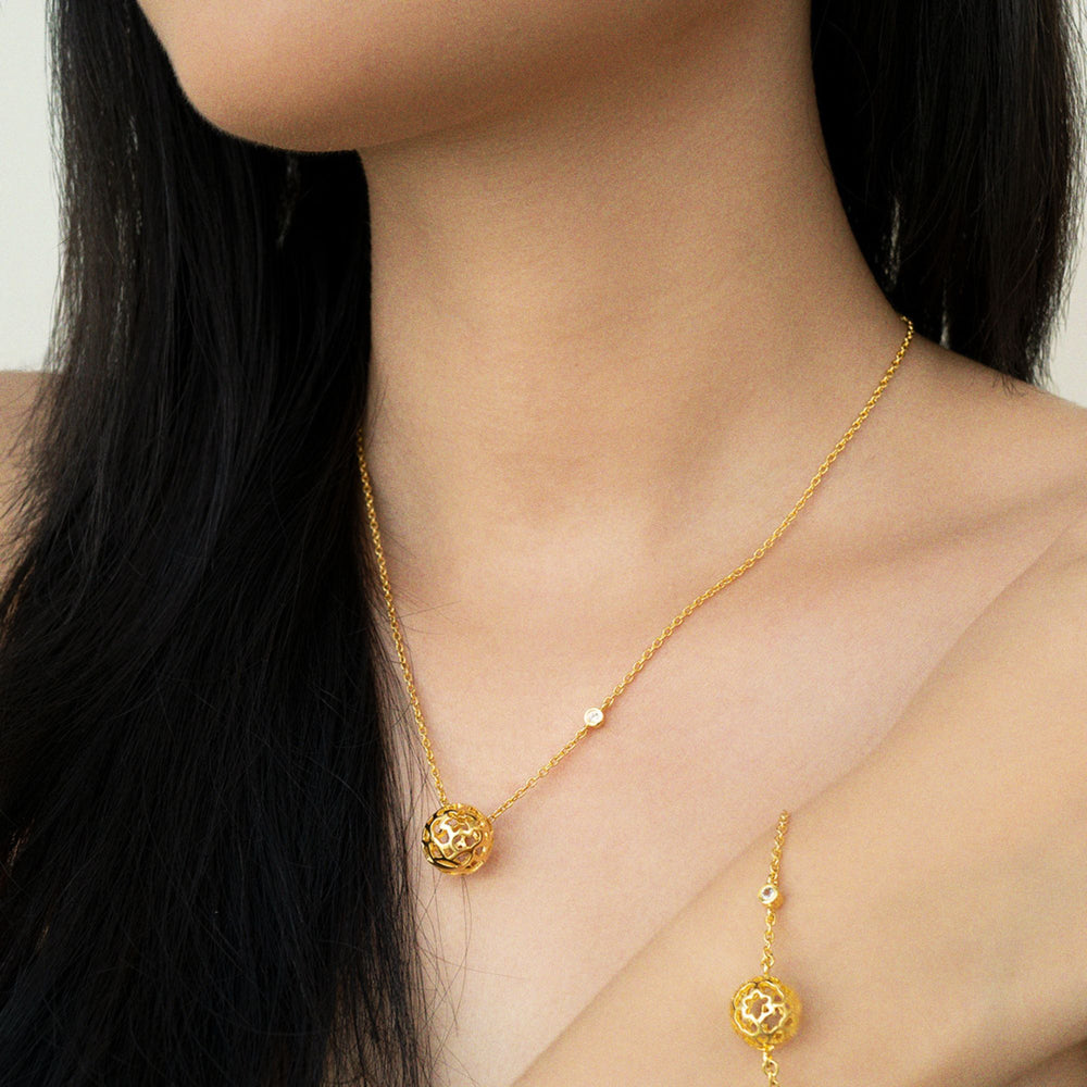 Risis Peranakan Spheres Large Necklace with White Topaz (G) - A golden necklace with a spherical, ornate pendant hangs around a person's neck, complemented by a smaller gemstone along its chain.
