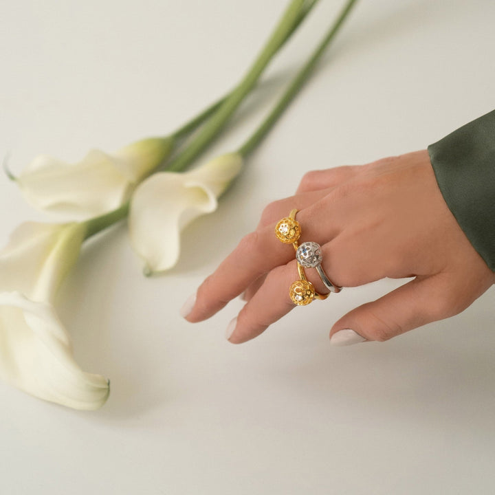 Risis Peranakan Spheres Large Ring with Blue Topaz (RH) - Three elegant rings with central gemstones displayed on a stone; two gold-banded rings with warm-toned jewels and one silver-banded with a cool-toned gem.