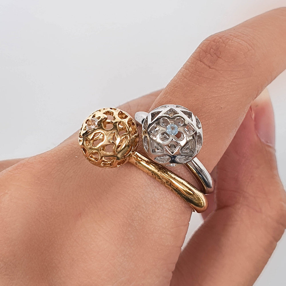 Risis Peranakan Spheres Large Ring with Blue Topaz (RH) - A hand adorned with two gold rings encrusted with sparkling gemstones rests elegantly, emphasizing the intricate designs and luxurious appeal of the jewelry.