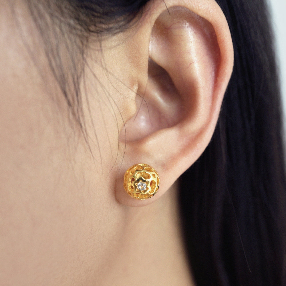 Risis Peranakan Spheres Pierced Earrings with White Topaz (G) - A golden earring, with intricate cut-out designs and a central jewel, adorns a person's earlobe, exhibiting elegance and craftsmanship.