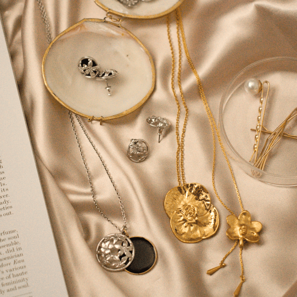 Risis Promesse Necklace (G) - Various necklaces and pendants with silver and gold tones, featuring floral and geometric designs, displayed elegantly on a satiny fabric surface.