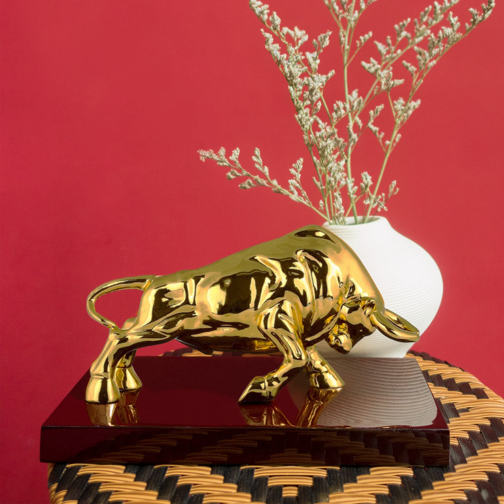 Risis Raging Bull - A reflective golden bull sculpture is positioned atop a dark-toned, textured surface, accompanied by a white vase with delicate beige flowers.