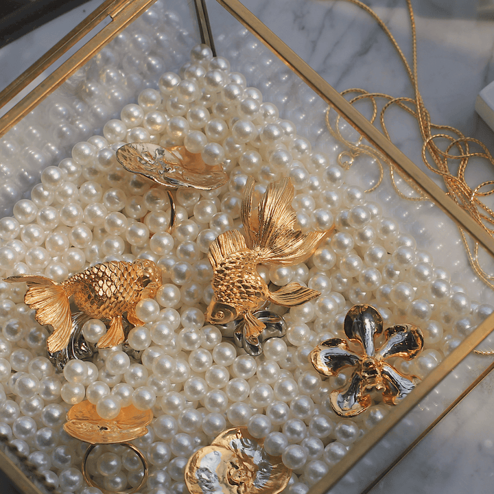 Risis Ranchu Goldfish Name Card Holder (Small) - Lustrous, white pearls fill a space, among which gold and copper-colored fish-shaped brooches, a florid ornament, and a sleek ring lie scattered, all exuding opulence and artistic craft.