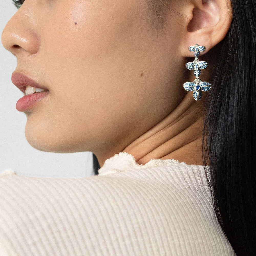 Risis Rare Orchid Earrings with Blue Sapphire - An elegant earring with a butterfly motif, featuring blue gemstones and silver settings, dangles gracefully from a woman's earlobe.