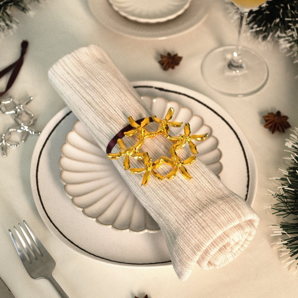 Risis Renantanda Sawadi Orchid Ornament (G) - A golden napkin ring with ribbon-like bows adorns a neatly folded cream napkin on a white plate, conveying elegance and festive cheer.