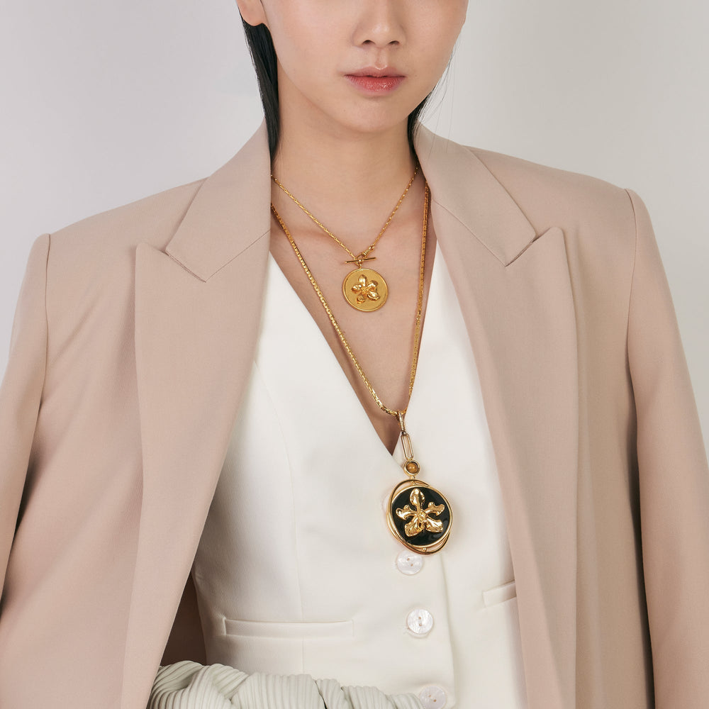 Risis X LINGWU Iconic Vanda Orchid Short Necklace - A person is wearing a beige blazer over a white top, adorned with two gold necklaces featuring pendants—one round with a flower motif, and one oval with a darker inlay.