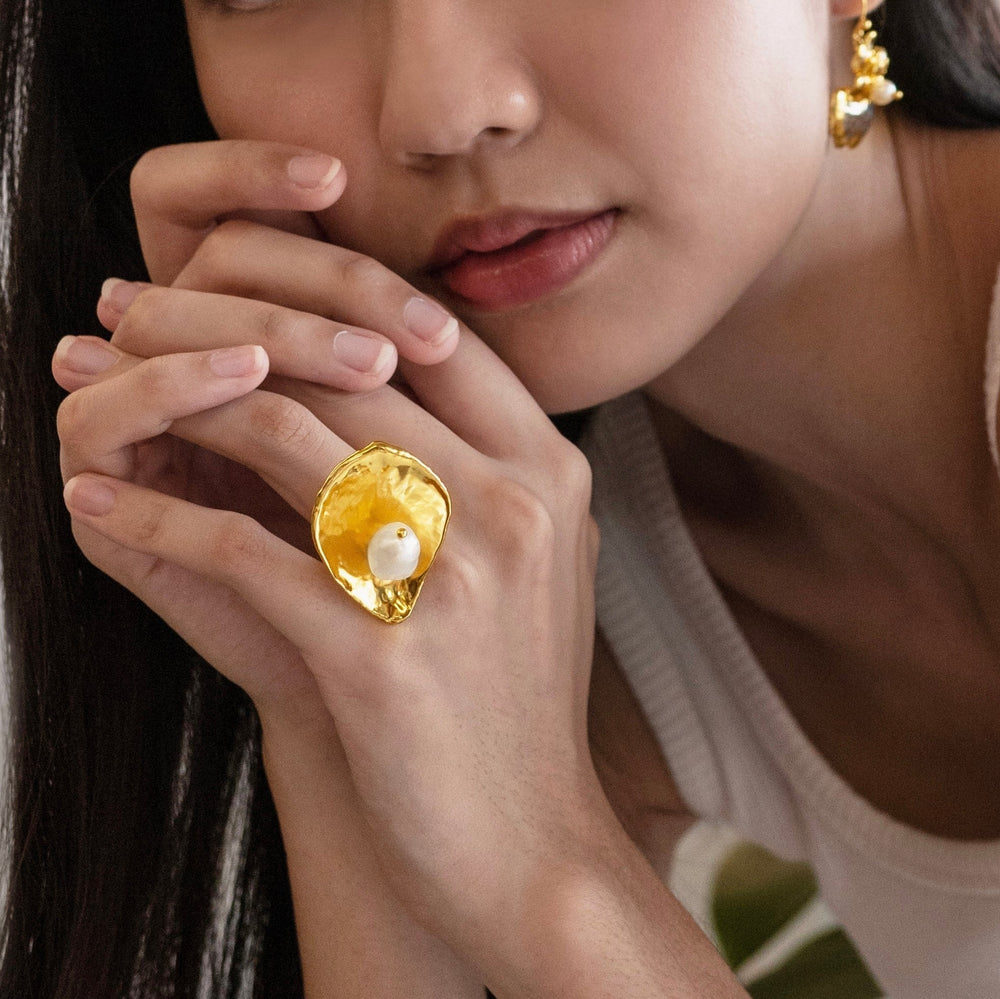 Risis Rose Petal Ring with Pearl - A large, lustrous golden ring with a textured surface and an embedded pearl is showcased on a person's finger, complemented by a gold earring with similar design elements.