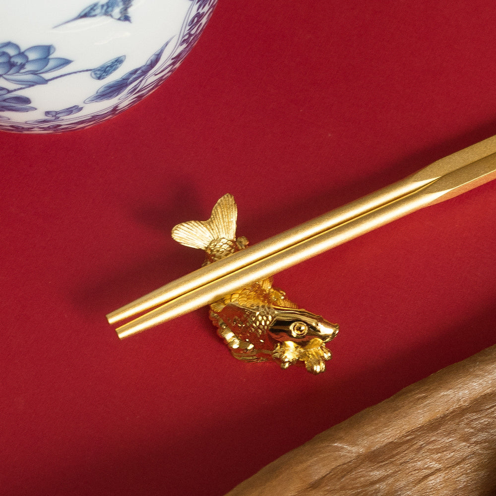 Risis Set of 6 Gold-Plated Chopsticks with Carp Rest - A golden chopstick rests atop a detailed fish-shaped holder with intricate scales and fins, against a vivid red backdrop. The fish features ornamental gold accents with a hint of red.