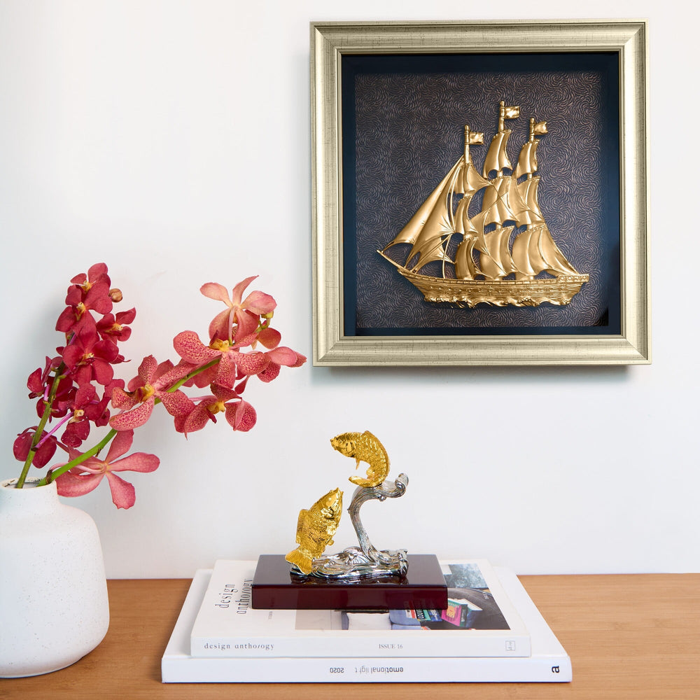 Risis Ship Frame - A golden ship sculpture with intricate sails is framed on a wall, and a metallic gold and silver fish sculpture stands on a table beside red orchids.