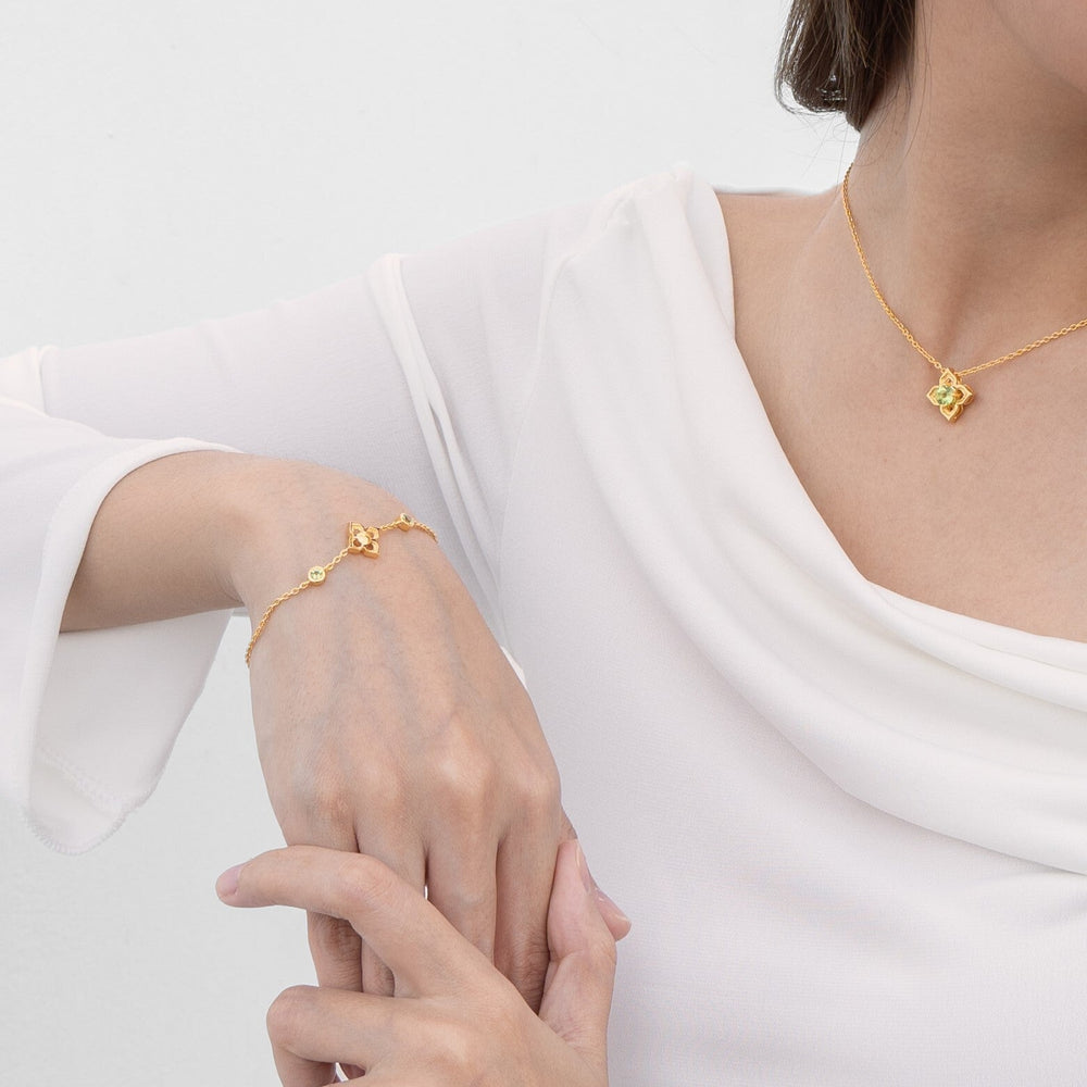 Risis Timeless Peranakan Bracelet With Peridot - A delicate gold bracelet with a tiny, ornate charm dangles from a woman's wrist, complementing the elegant gold necklace with a similar pendant she wears.
