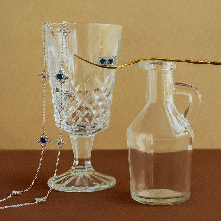 Risis Timeless Peranakan II Necklace (RH) with London Blue Topaz - A crystal glass and a transparent bottle are adorned with delicate jewelry featuring sparkling chains and deep blue gemstones emphasizing elegance and refinement.