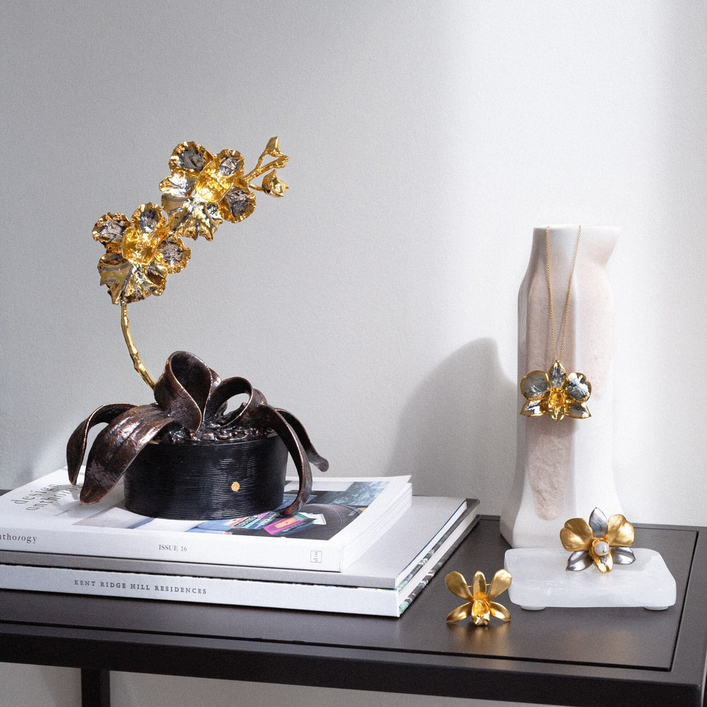 Risis Vanda Miss Joaquim Orchid In Pot - A golden sculpture of leaves and flowers emerges from a dark, ribbon-like vase set on books, paired with similarly styled jewelry displayed beside a white form.