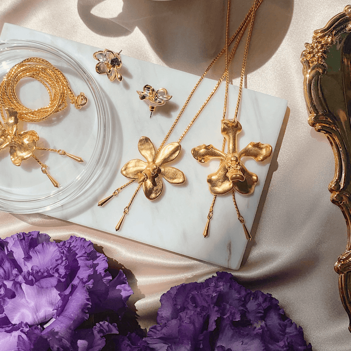 Risis Vanda Wee Kim Lian Orchid Slider Necklace (G) - Golden jewelry with floral motifs, including necklaces and earrings, showcased amidst soft lighting, enhancing their lustrous appeal and delicate craftsmanship.