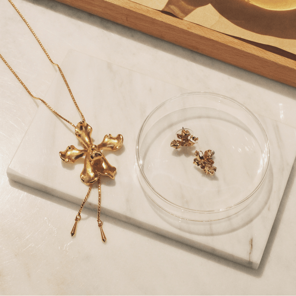 Risis Vanda Wee Kim Lian Orchid Slider Necklace (G) - A gold, flower-shaped pendant with a textured surface is displayed, with two golden chains draped elegantly. Nearby, small, intricately designed gold earrings rest on a clear disc.