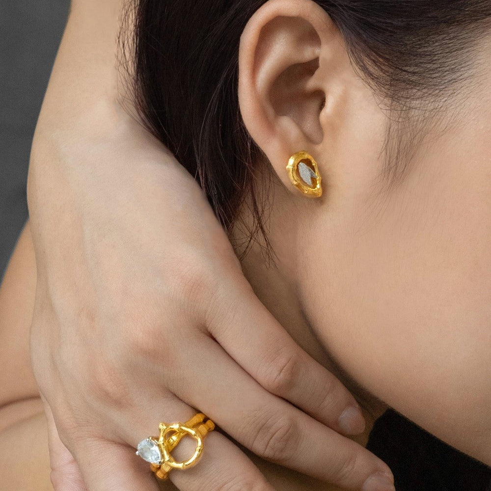 Risis Vintage Bamboo Earrings with White Topaz - A golden earring with an oblong jewel dangles from an ear, while a hand adorned with a gold ring featuring a clear gemstone rests upon a shoulder.