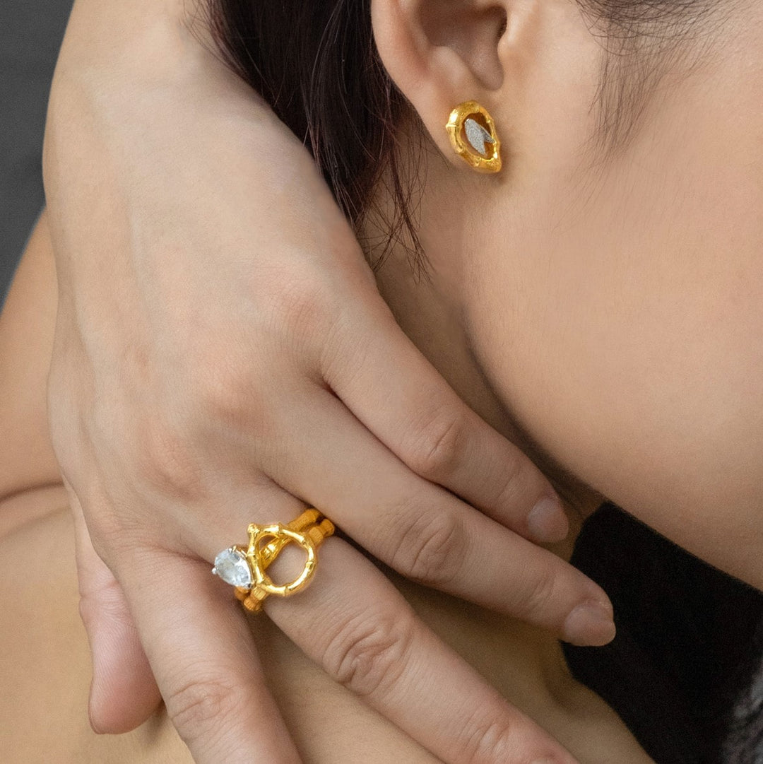 Risis Vintage Bamboo Ring with White Topaz - A person is wearing a golden earring with an oval clear gemstone and a matching golden ring with a similar stone, showcasing jewelry elegance.