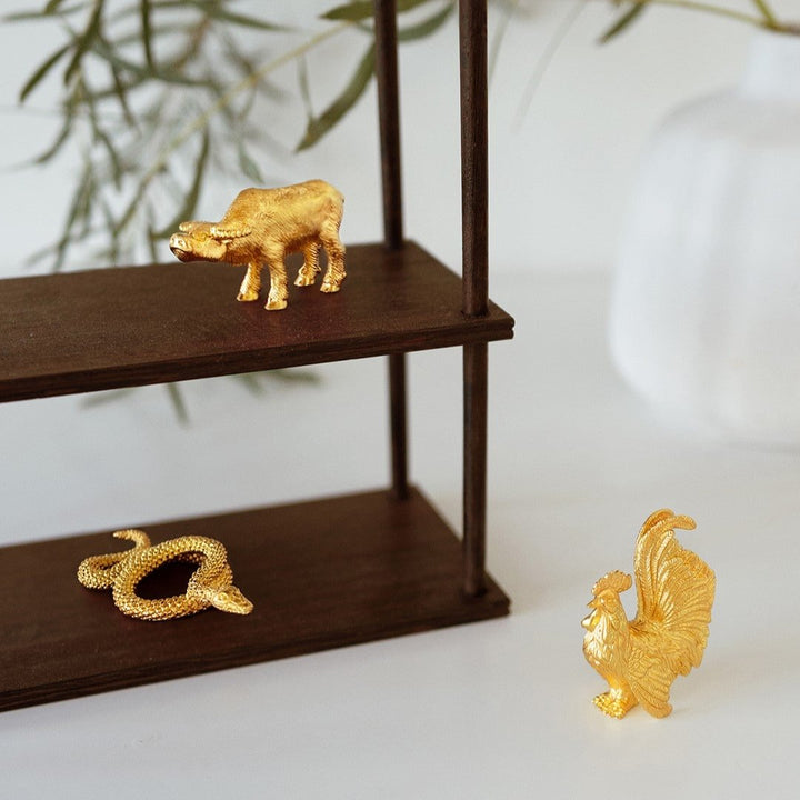 Risis Zodiac Snake Figurine - A golden hippopotamus, snake, and rooster figurine rest on dark wooden shelves, shimmering with intricate textures and a lifelike appearance.