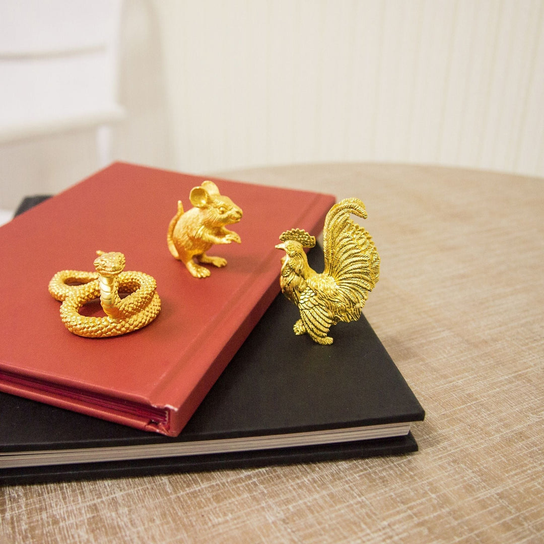 Risis Zodiac Snake Figurine - Gold animal figurines—a mouse, a snake, and a rooster—adorn two stacked books, showcasing intricacy and lustrous texture.